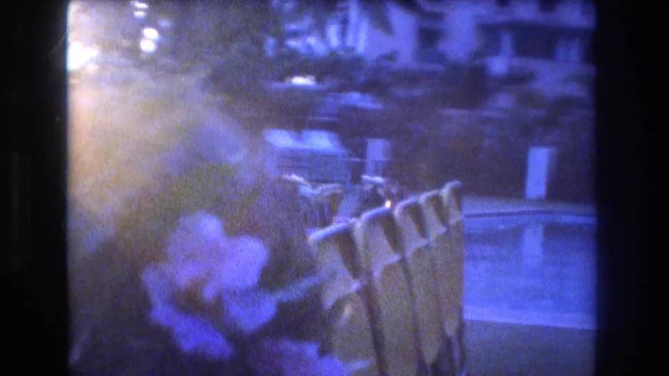 1977 Honolulu home-movie frame showing a blue twilight pool courtyard with lounge chairs, tropical flowers, foliage, and multi-story buildings beyond the pool.