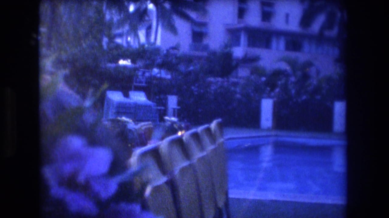 Poolside chairs beside a courtyard pool with tropical plants and buildings behind them.