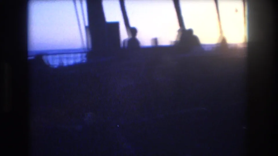1977 Honolulu 8mm frame with indistinct silhouetted figures near a deck or railing, set against a bright dusk sky that may overlook the coast.