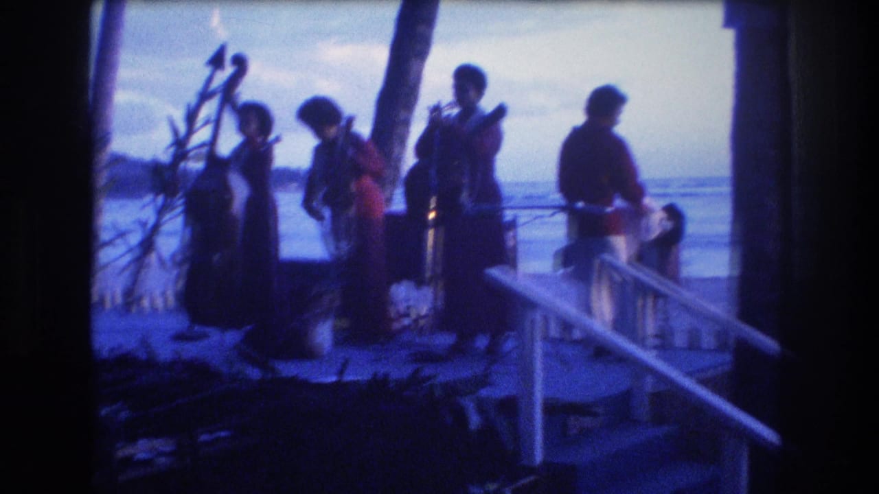 Women in long dresses stand with instruments near a railing overlooking the ocean.