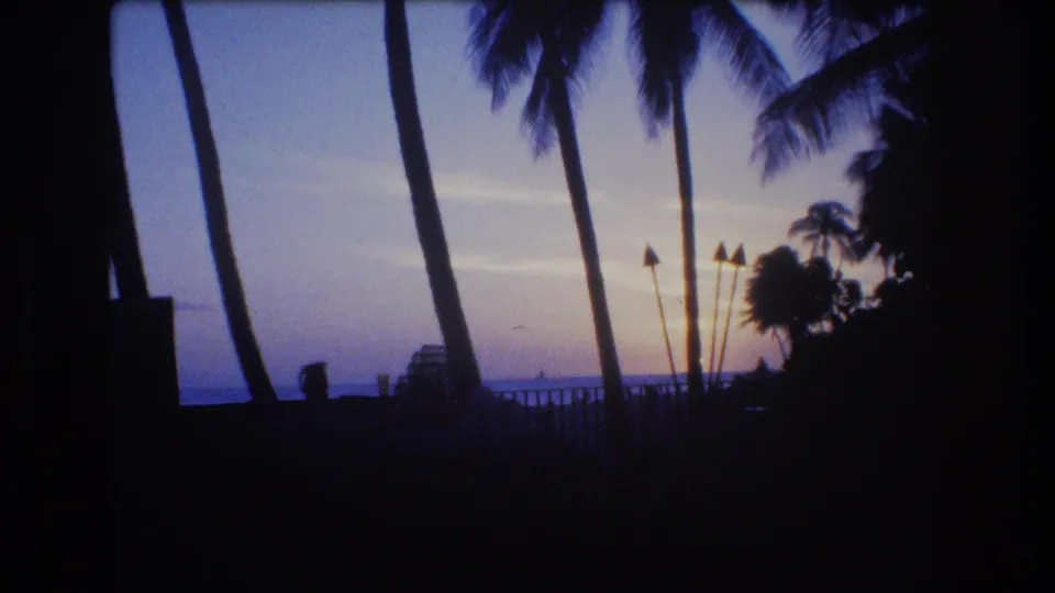 1977 Honolulu home-movie frame showing a quiet sunset view through palm trees, with a shadowed patio or balcony area in the foreground and ocean beyond.