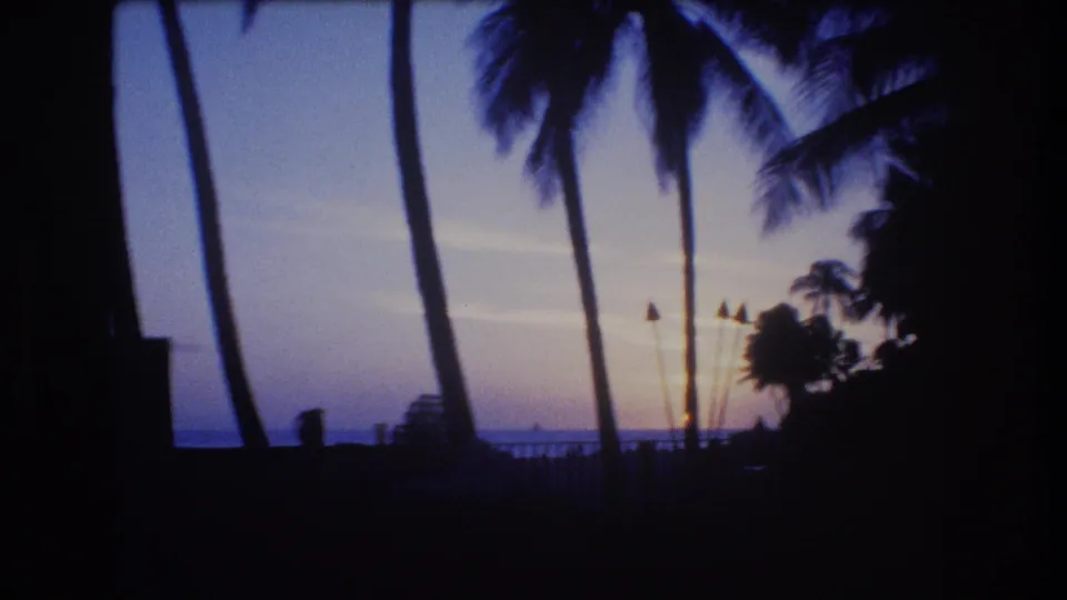 Final 1977 Honolulu sunset frame with palm silhouettes, torch-like poles, a dark railing, and the ocean horizon under a fading blue and orange sky.