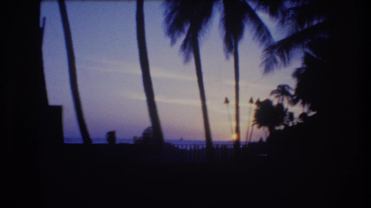 1977 Honolulu or Oahu 8mm frame showing palm trees, tiki torches, a railing, and the sun low over the ocean horizon at twilight.