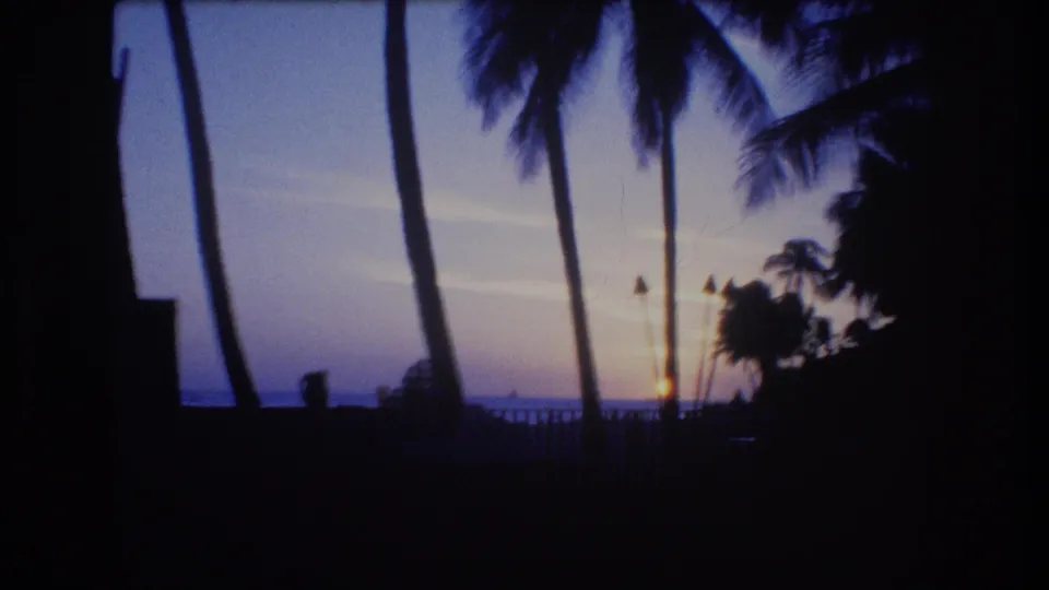 1977 Honolulu or Oahu 8mm frame showing palm trees, tiki torches, a railing, and the sun low over the ocean horizon at twilight.