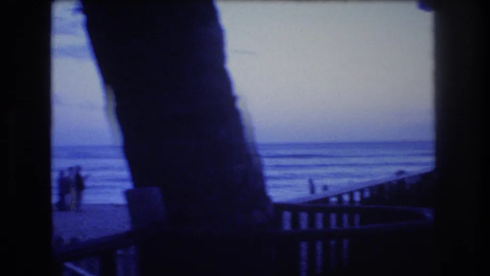 1977 Honolulu or Oahu frame with silhouetted people near the water, calm waves, and large dark palm trunks or posts in the foreground.