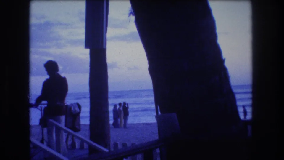 1977 Honolulu or Oahu home-movie frame showing a silhouetted adult beside a railing, with distant figures, ocean waves, and dark palm trunks at dusk.