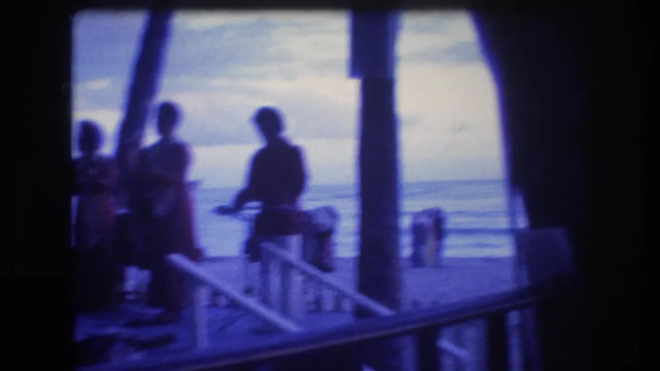 1977 Honolulu or Oahu frame showing three silhouetted adults near a deck railing, with palm trunks, dusk sky, and the ocean in the background.