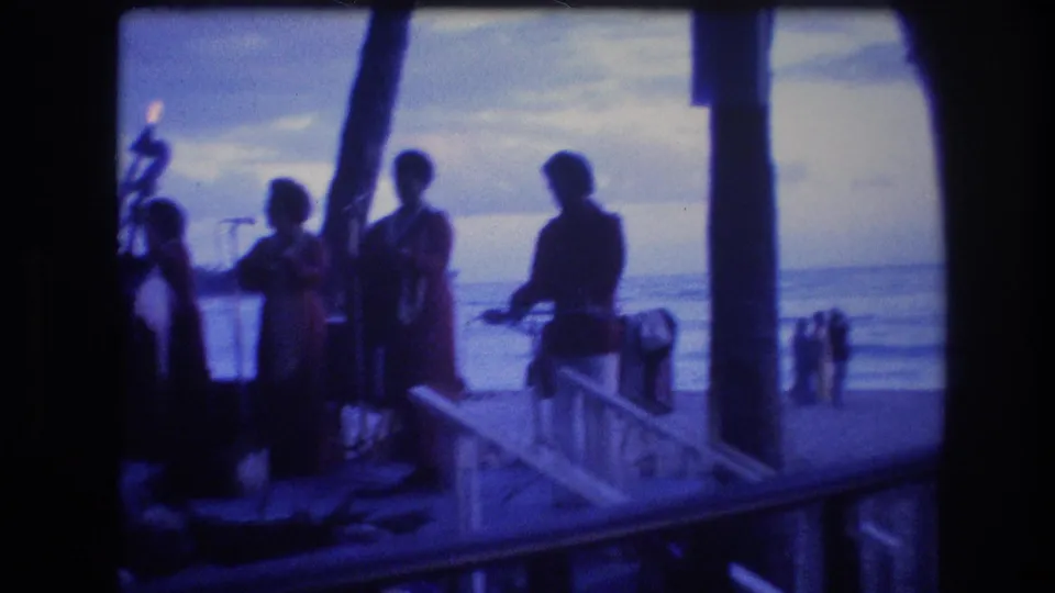 Silhouetted performers appear on a beachside pier or terrace in 1977 Honolulu, with ocean water and a dim evening sky behind them.