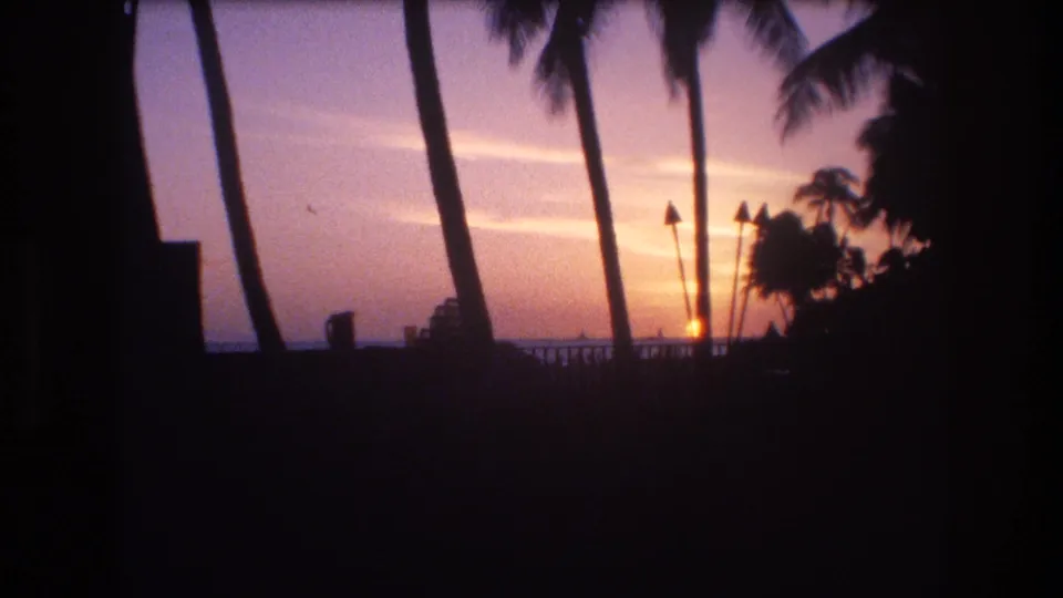 1977 Honolulu, Oahu 8mm frame showing palm trunks in silhouette against a warm sunset sky over the ocean, with a dark railing and foreground shadows.