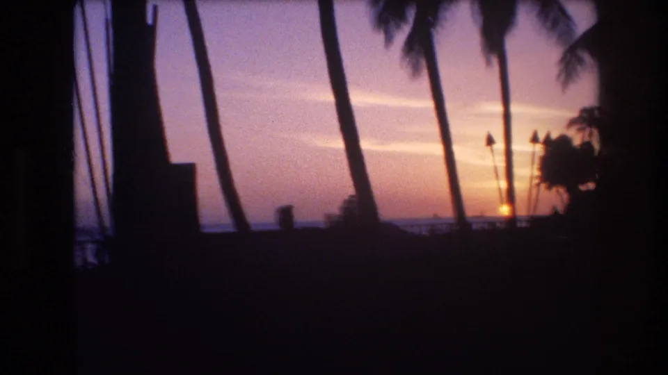 1977 Honolulu, Oahu 8mm frame with a low orange sun near the ocean horizon, silhouetted palms, and dark foreground structure along the edge of the view.