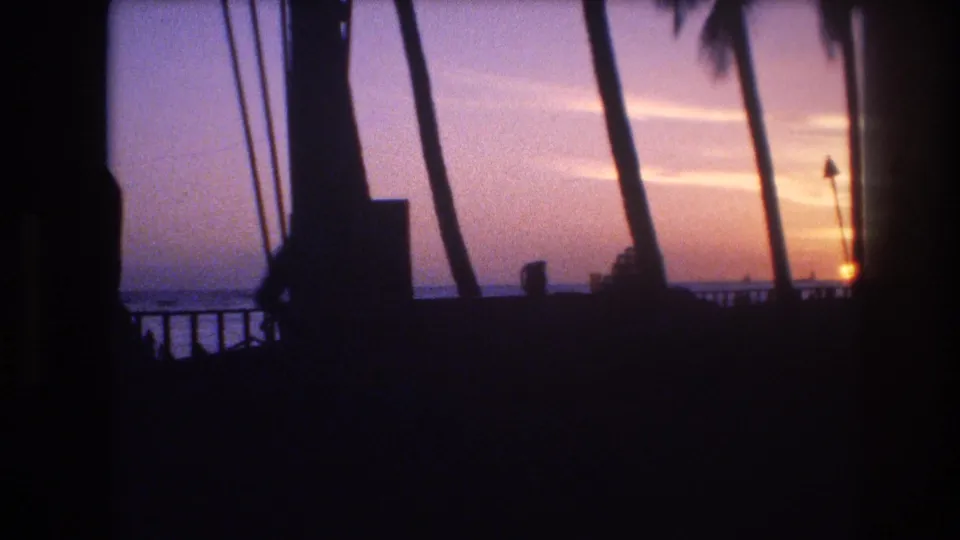 1977 Honolulu, Oahu 8mm frame showing a soft sunset sky over water, partly obscured by palm trunks, a railing, and dark silhouetted foreground forms.