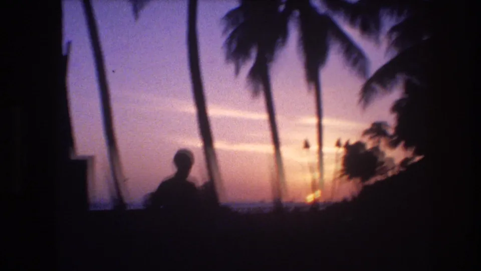1977 Honolulu, Oahu 8mm frame with a silhouetted person in the foreground, palm trees overhead, and a warm sunset over the ocean beyond.
