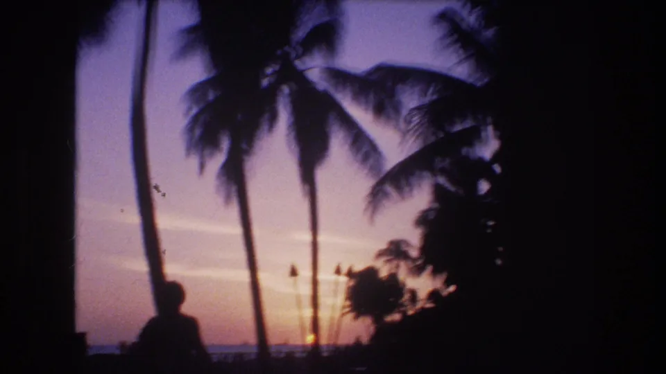 A 1977 Honolulu home-movie frame appears to show a silhouetted person walking near palm trees as the sun sets over the shoreline.