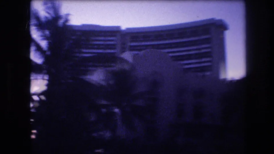 A softly focused 1977 Honolulu frame shows palm foliage partly obscuring a high-rise resort building against a blue-purple twilight sky.