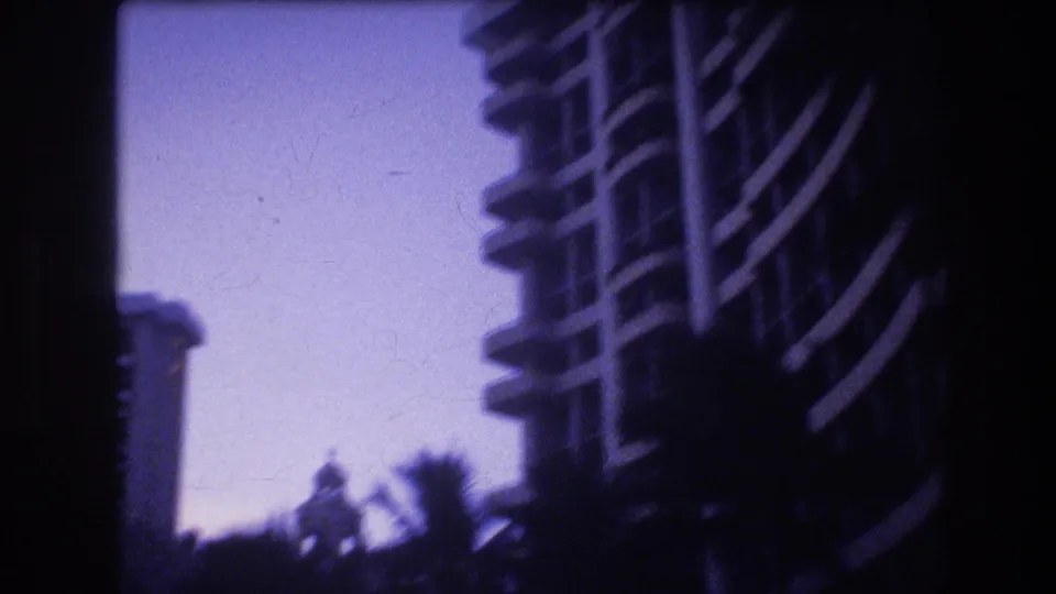 A 1977 Honolulu home-movie view shows a tall hotel or resort building with repeating balconies partly hidden by dark tropical foliage.