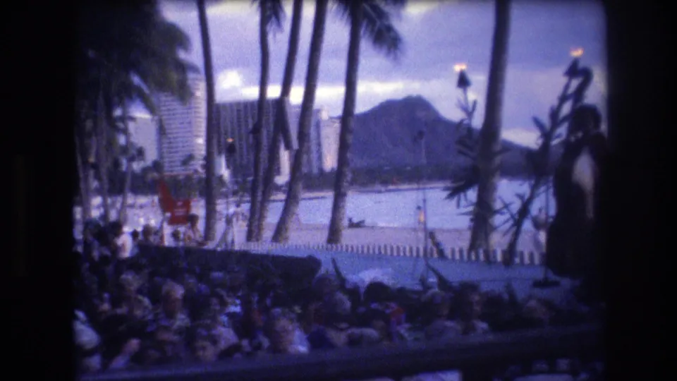 1977 Honolulu home-movie frame with spectators in the foreground, a small outdoor performance area, tiki torches, palms, shoreline water, hotels, and Diamond Head in the distance.
