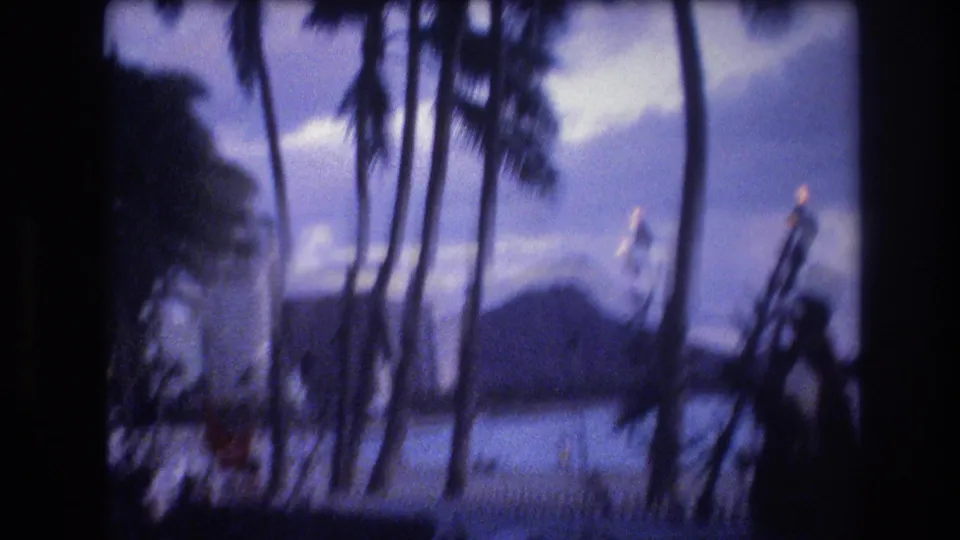 Soft 8mm Honolulu frame from 1977 showing palm trunks and dark foreground silhouettes around a beachside entertainment setting, with water and Diamond Head visible behind.
