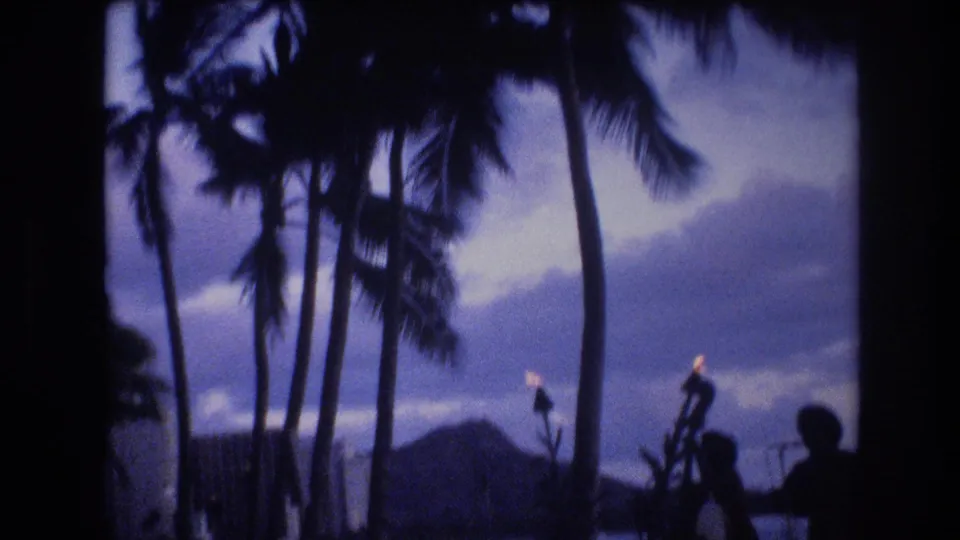 1977 Honolulu 8mm frame with silhouetted palms, torchlight, shoreline water, and Diamond Head in the background, suggesting the end of a brief beachside entertainment shot.