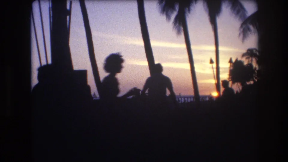 1977 Honolulu, Oahu home-movie frame with dark seated figures near palm trunks, a low sun, torch shapes, and waterfront sky at dusk.