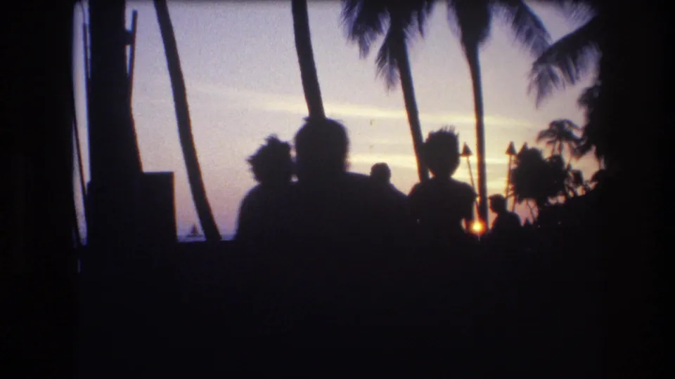 1977 Honolulu, Oahu frame showing a small group of silhouetted figures beneath palm trees, with the setting sun and waterfront behind them.