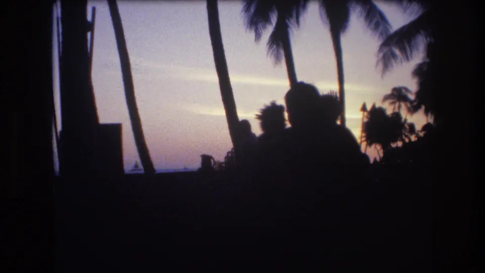 1977 Honolulu, Oahu home-movie frame with dark silhouettes of people under palm trees looking toward a fading sunset over the ocean.