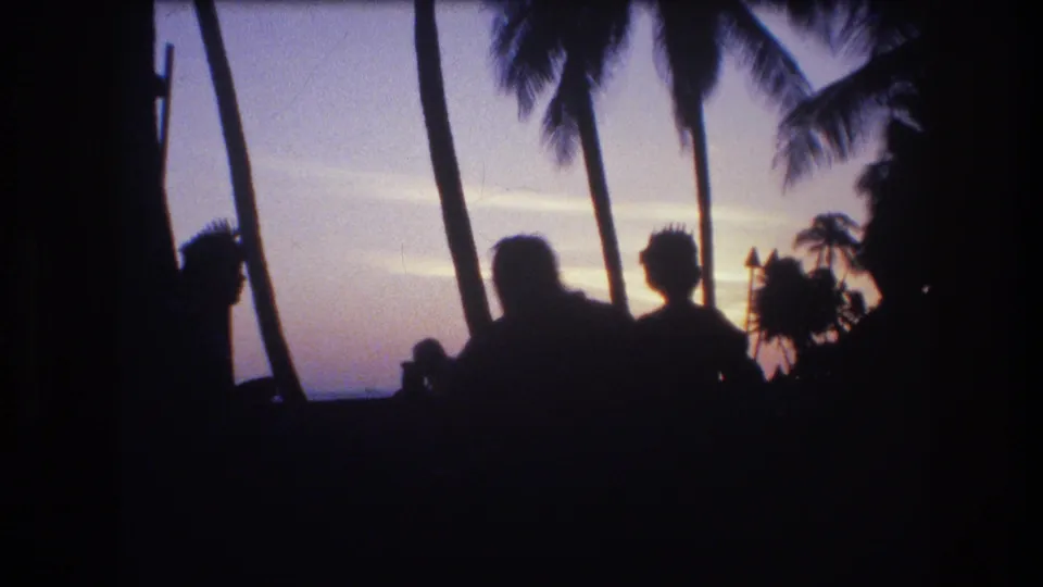 A 1977 Honolulu, Oahu sunset frame shows two prominent dark human silhouettes near palm trees, with the beach and ocean horizon beyond.
