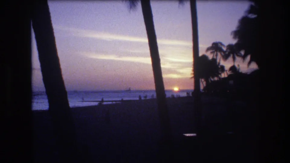1977 Honolulu 8mm frame showing the sun near the ocean horizon, silhouetted palms, purple evening sky, and small figures along the beach.