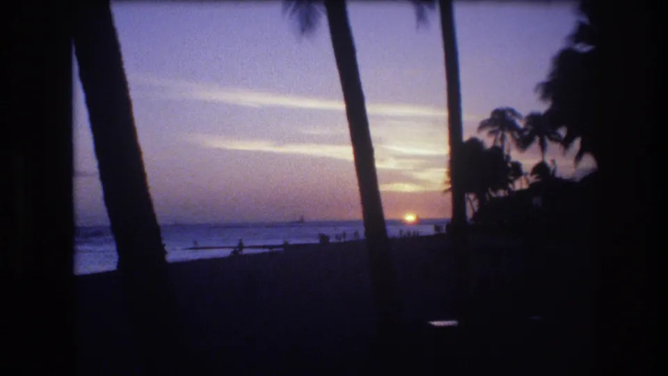 1977 Honolulu beach view with the sun close to the horizon, dark palm silhouettes, surf, and distant beachgoers at dusk.