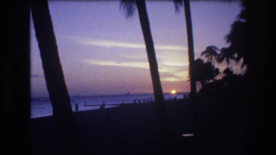 1977 Honolulu 8mm frame with a warm sunset, purple sky, silhouetted palms, and small people near the shoreline.