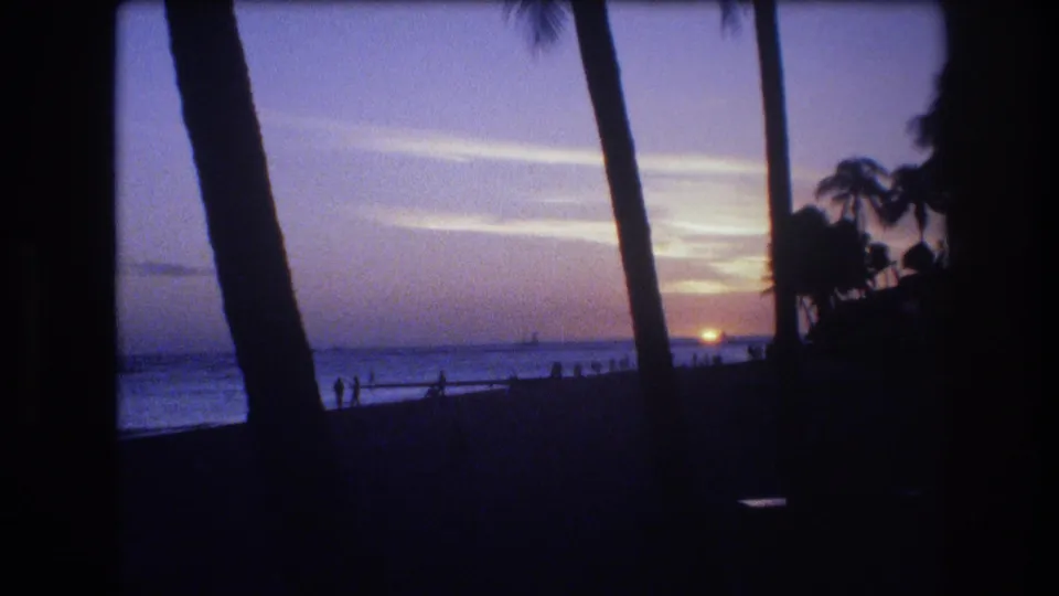1977 Honolulu beach sunset frame showing distant people near the surf, tall palm silhouettes, and a fading orange sun over the water.