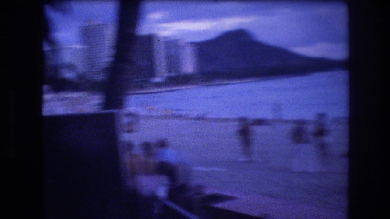 Blue-tinted 1977 Honolulu home-movie frame showing shoreline and ocean with Diamond Head visible in the distance.