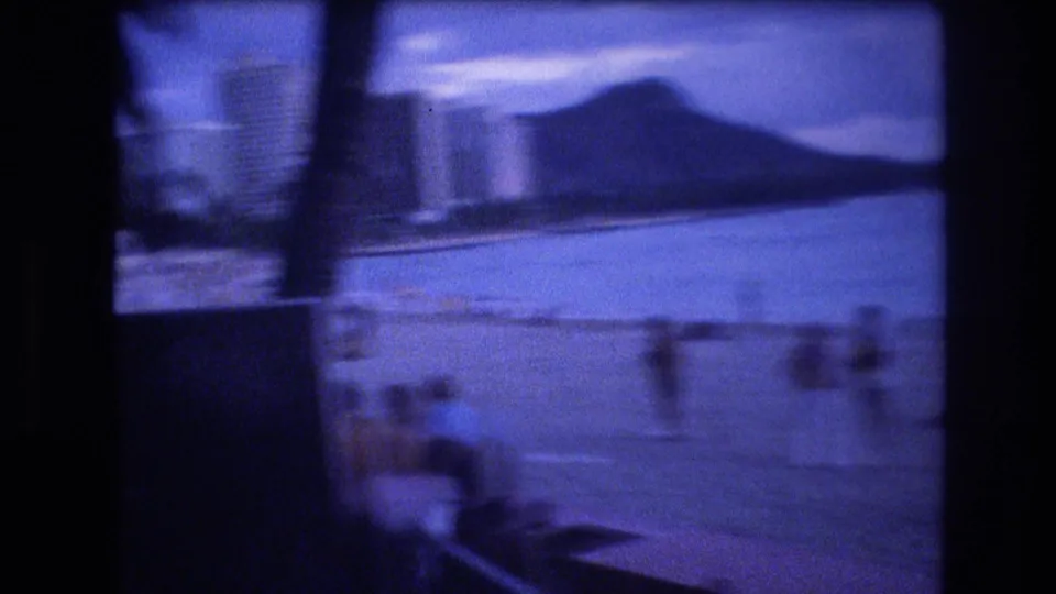 Blue-tinted 1977 Honolulu home-movie frame showing shoreline and ocean with Diamond Head visible in the distance.