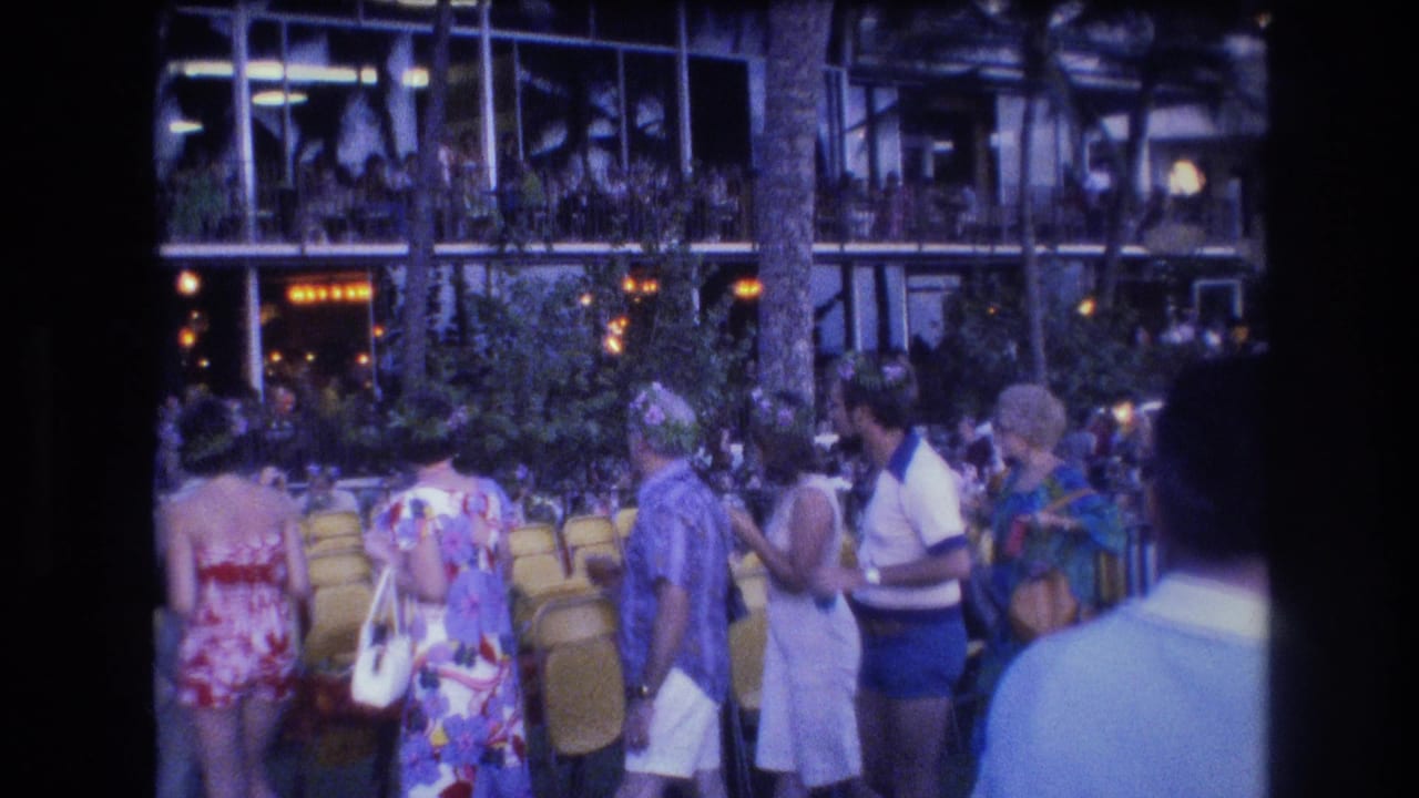 Handheld 1977 Honolulu home-movie frame showing tourists in swimwear, floral dresses, and casual resort clothing beside yellow chairs and palm trunks.