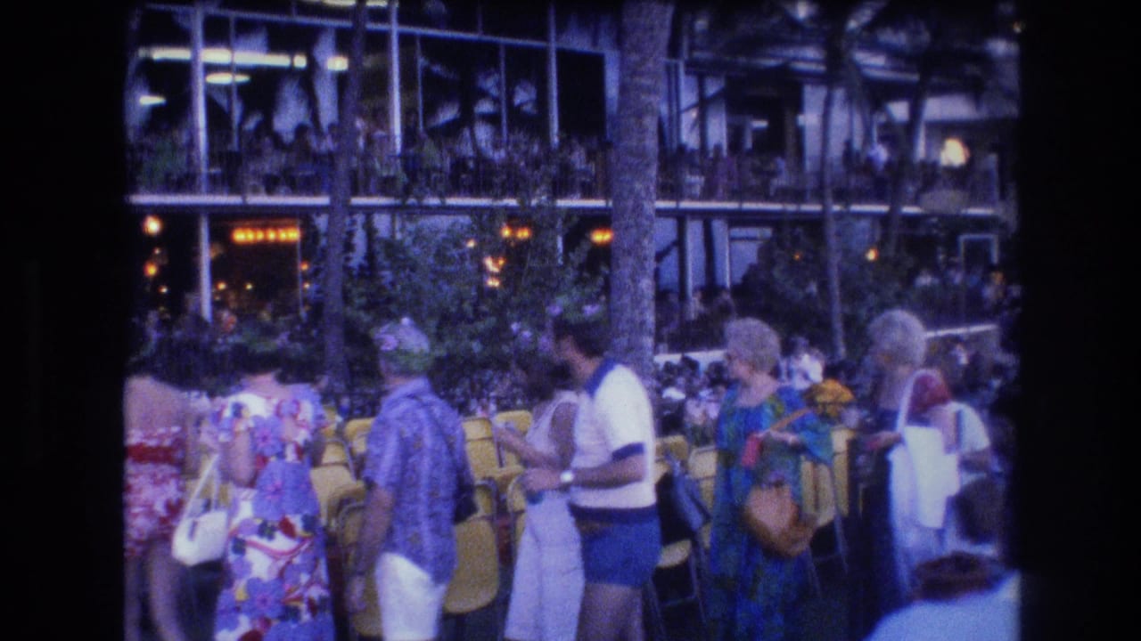 1977 Honolulu frame of a casual tourist crowd near yellow resort chairs, palm landscaping, and illuminated glass-fronted architecture.