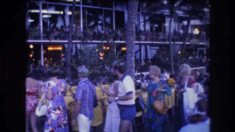 1977 Honolulu frame of a casual tourist crowd near yellow resort chairs, palm landscaping, and illuminated glass-fronted architecture.