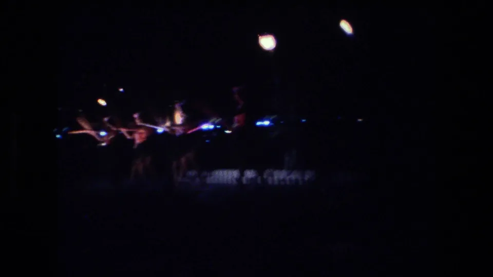 Distant 1977 Honolulu home-movie view of a nighttime Hawaiian torch dance, with performers barely visible under dark exposure and bright flame points.
