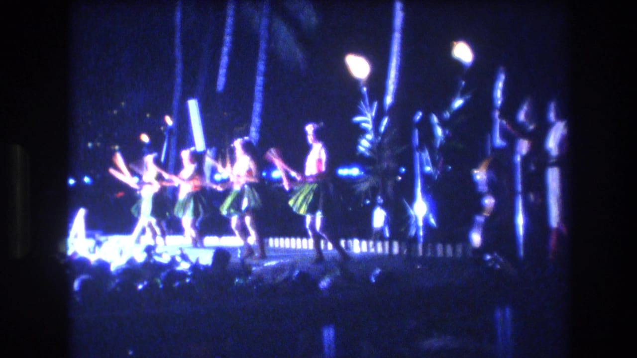 1977 Honolulu nighttime performance frame showing several Hawaiian dancers on a bright stage with flaming torches, blue lights, and dark palm-like shapes behind them.