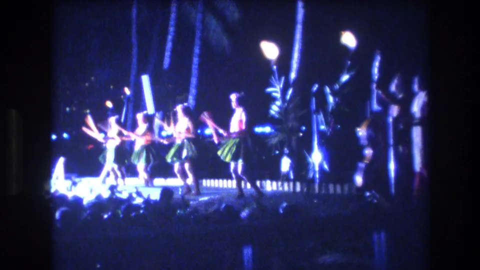 1977 Honolulu nighttime performance frame showing several Hawaiian dancers on a bright stage with flaming torches, blue lights, and dark palm-like shapes behind them.