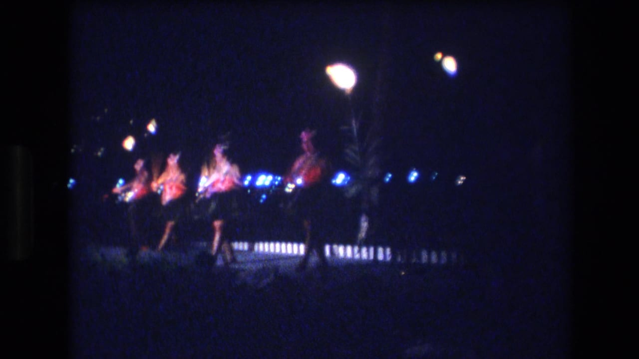 1977 Honolulu home-movie frame of dancers in colorful tops and skirted costumes moving across a dim outdoor stage beneath glowing lamps and blue light.