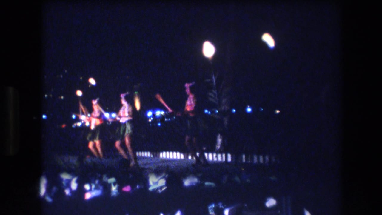 1977 Honolulu nighttime dance-performance frame with performers holding torches near blue stage lights, a low railing, and shimmering reflections in the foreground.