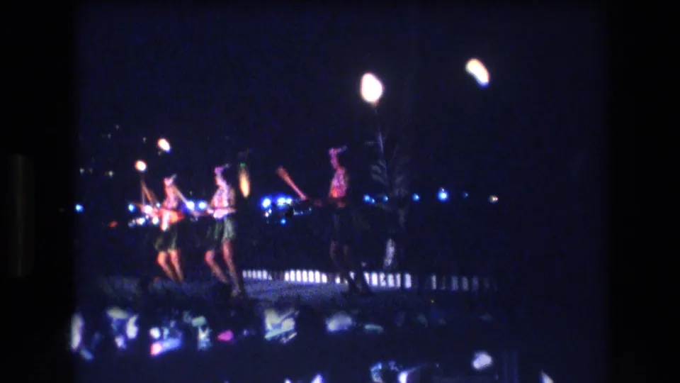 1977 Honolulu nighttime dance-performance frame with performers holding torches near blue stage lights, a low railing, and shimmering reflections in the foreground.