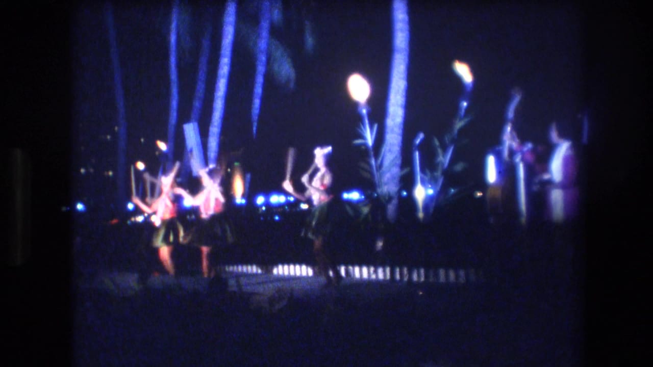 1977 Honolulu 8mm frame showing distant hula dancers in grass-style skirts performing at night under glowing lights with palm trunks nearby.