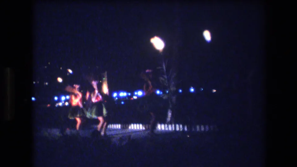 1977 Honolulu frame with hula dancers moving through a low-light outdoor performance area, backed by blue bulbs and tall glowing stage lamps.