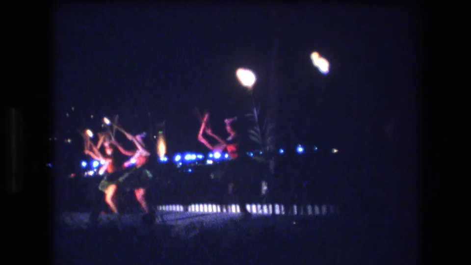 1977 Honolulu 8mm still with foreground hula dancers raising their arms during a night performance, with bright lamps and blue stage lights in the background.