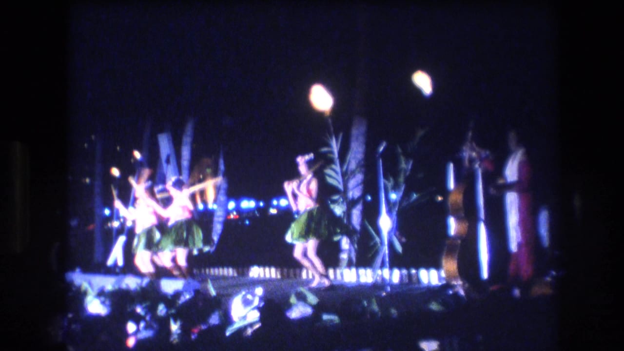 1977 Honolulu home-movie frame showing dancers in grass skirts moving under torchlight on a dark outdoor luau-style stage.
