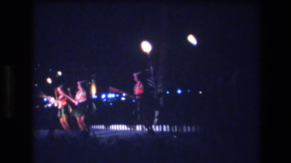 1977 Honolulu frame with dancers in grass skirts grouped near tall torches, blue lights, and a dark nighttime backdrop.
