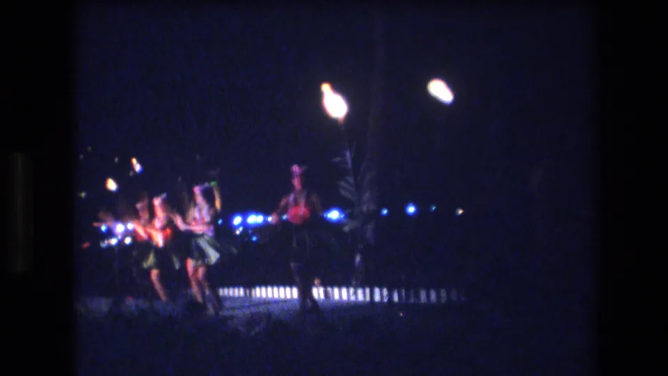 1977 Honolulu 8mm frame showing luau-style dancers crossing a dim outdoor stage with torch flames and blue lights in the background.