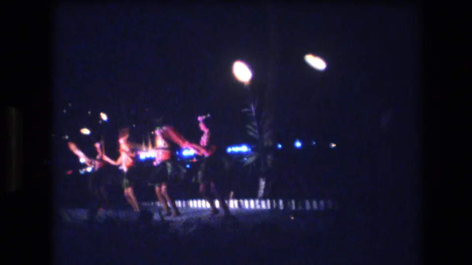 1977 Honolulu home-movie frame showing dancers in grass skirts performing in a loose line beneath glowing torches on a dark stage.