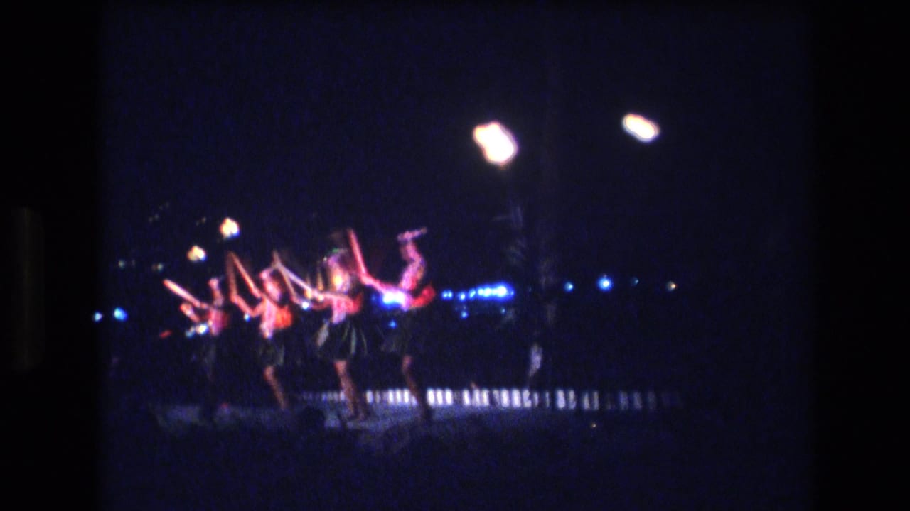 Women perform a nighttime line dance under bright lamps and blue accent lights.