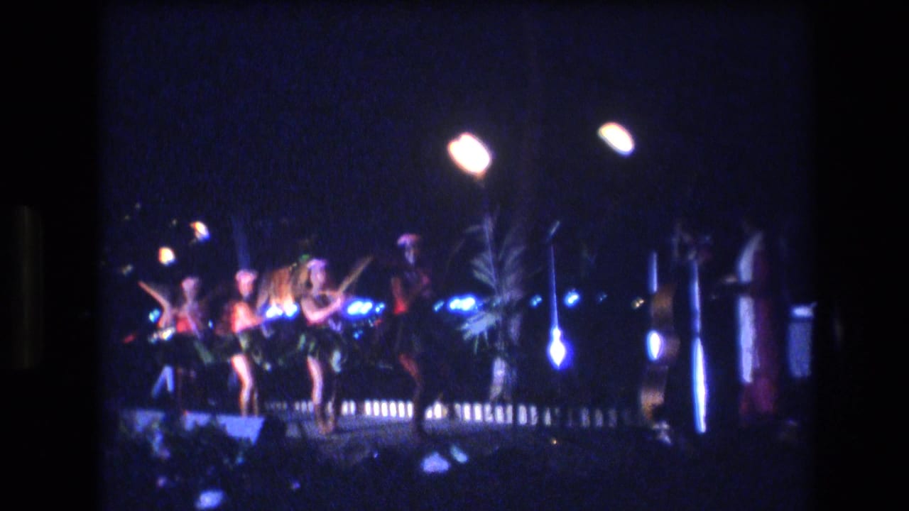 A line of dancers moves across a lit outdoor stage area at night.